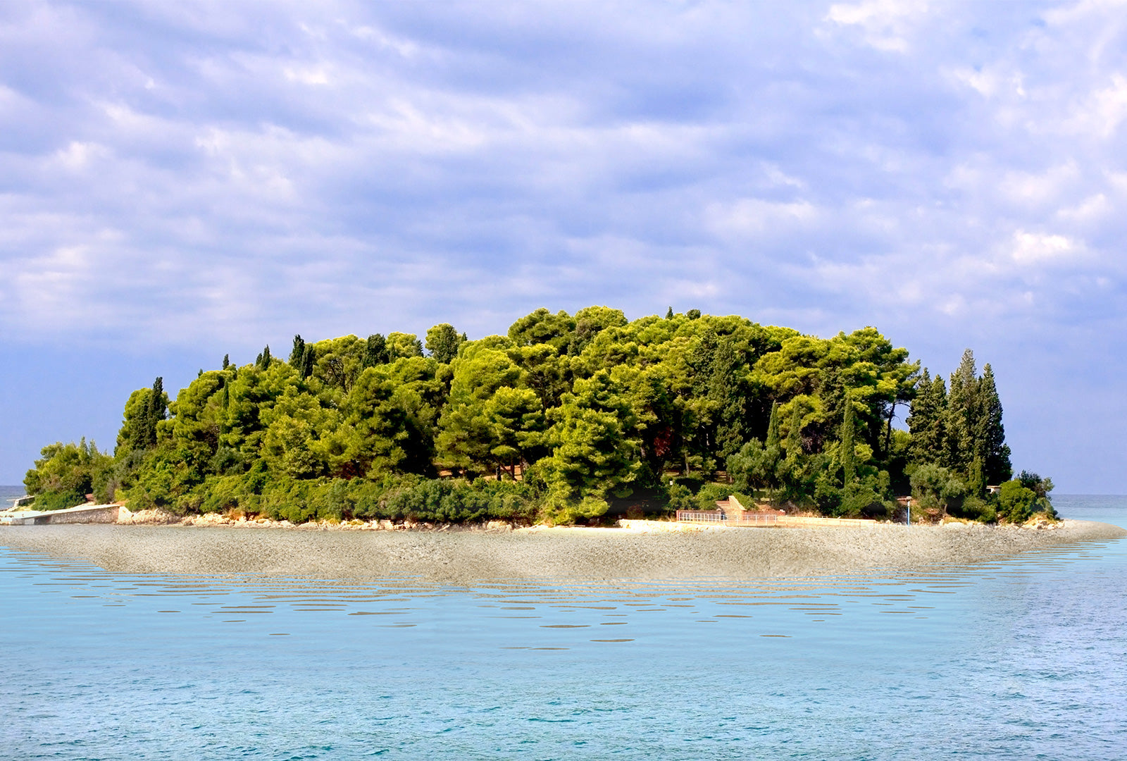 Island with lush greenery surrounded by clear blue water under a partly cloudy sky